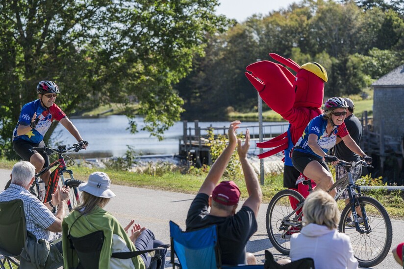two cyclists riding along a river with people cheering them on in the foreground and a lobster mascot in the background